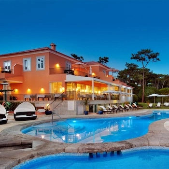 Vista exterior de hotel e clínica ao entardecer com piscina iluminada, espreguiçadeiras e terraço